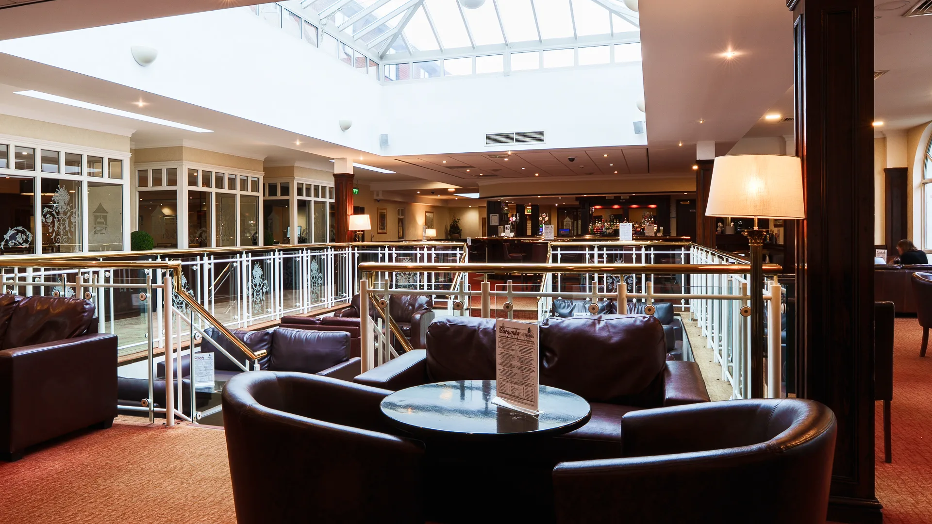A spacious hotel lounge with leather armchairs and tables arranged around an open balcony area. Large skylights and windows fill the space with natural light, while a bar area is visible in the background beneath warm interior lighting.